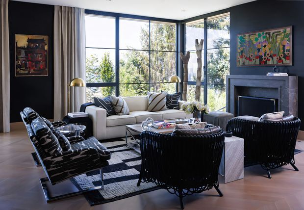 A sophisticated contemporary living room with expansive black-framed corner glazing, a white sofa, black leather lounge chairs, sculptural black woven armchairs, brass floor lamps, patterned cushions, and a dark stone fireplace wall. Timber flooring, layered rugs, and colourful artwork add warmth and visual interest, while the large windows bring in abundant natural light and views of mature trees.