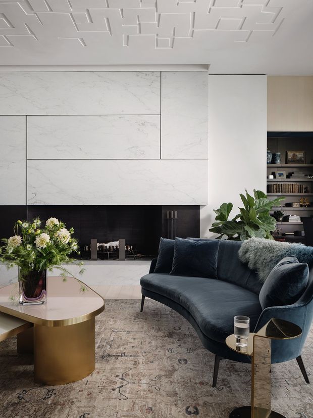 A refined contemporary living room with a deep navy curved sofa, curved brass coffee table, patterned neutral rug, and a dark side table. The focal wall combines large white marble-look panels with a dark fireplace surround and integrated shelving. A sculptural geometric white ceiling feature adds architectural interest, while plants and flowers soften the space.