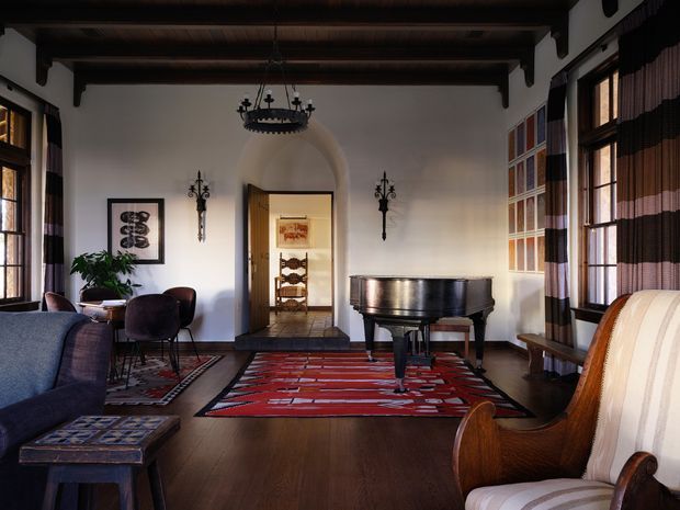A richly furnished, traditional living room with dark exposed ceiling beams, polished dark timber flooring, white plaster walls, and a grand piano as the focal point. The room features a patterned red area rug, upholstered armchairs and sofa, tall timber-framed windows with striped curtains, decorative wall sconces, and a central doorway leading to another room. The atmosphere is refined, warm, and slightly dramatic, with strong historical and eclectic detailing.