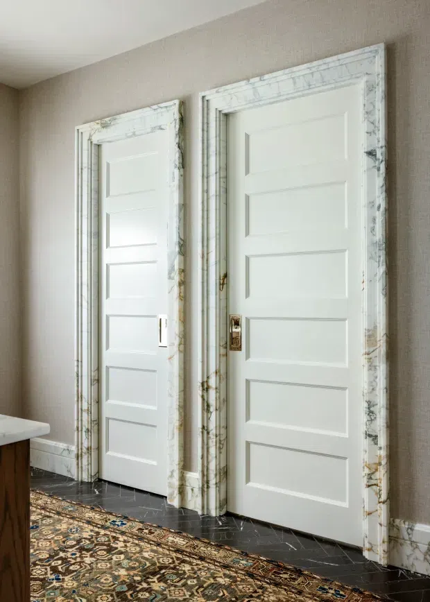 Interior view of two white paneled doors with ornate veined marble surrounds, set against textured taupe wallcovering. A dark marble floor, patterned oriental rug, white baseboards, and brass door hardware create a refined, traditional hallway or entry setting.