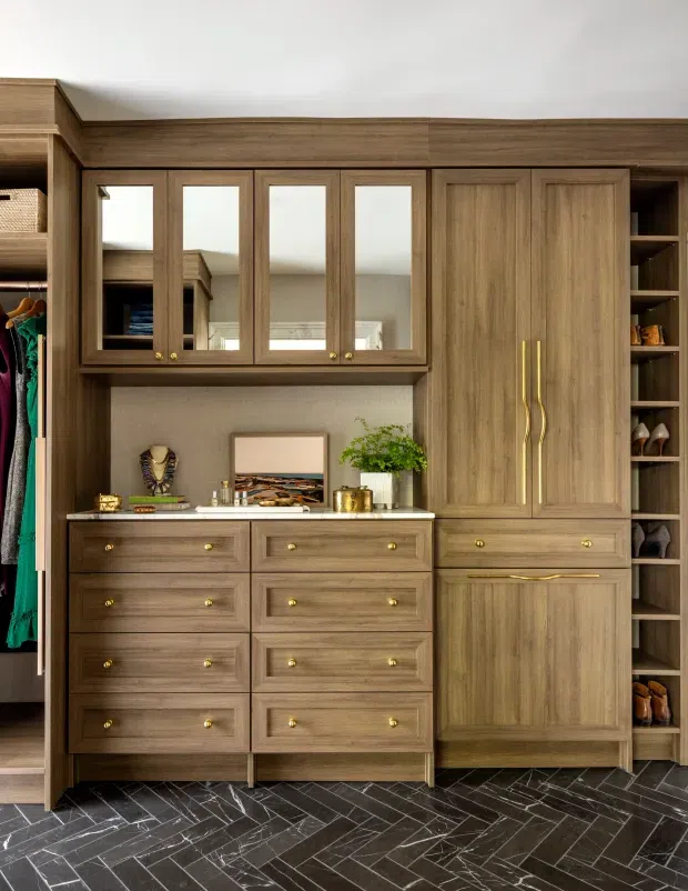 A built-in dressing room or walk-in wardrobe with extensive warm timber cabinetry, mirrored upper doors, drawers, tall wardrobe storage, open shelving, and brass hardware. Clothing and shoes are visible at the sides. The floor is finished in dark grey herringbone tile, with a neutral wall and a small styled countertop display.