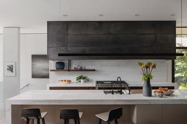 Contemporary open-plan kitchen with a large white marble-look island and integrated seating, dark timber or metal feature cabinetry spanning the wall, a long black rangehood, and a matching marble-look splashback. Recessed ceiling lights, minimalist decor, and large glazing create a refined, modern interior with views to greenery outside.