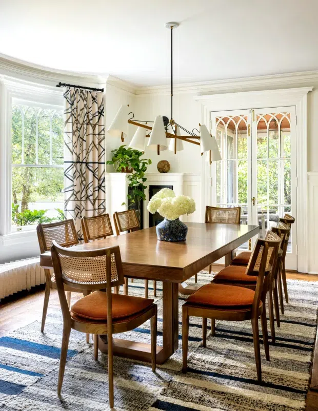 Bright, elegant dining room with a large rectangular timber dining table surrounded by woven-back wooden chairs with rust-coloured upholstered seats. The room features white walls with decorative moulding, tall multi-pane windows, glazed French doors with Gothic-inspired geometric detailing, a fireplace, patterned curtains, a sculptural multi-arm pendant light, and a large patterned area rug. Lush greenery is visible outside the windows.