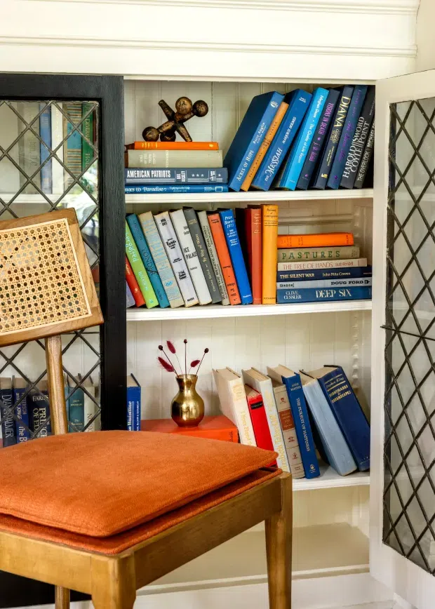 A cozy, traditional reading nook featuring a built-in white bookshelf filled with colourful books, framed by dark leaded-glass cabinet doors. A wooden chair with a deep rust-orange upholstered cushion occupies the foreground, while a small brass vase and decorative objects add warmth. White vertical wall panelling and classic trim complete the setting.