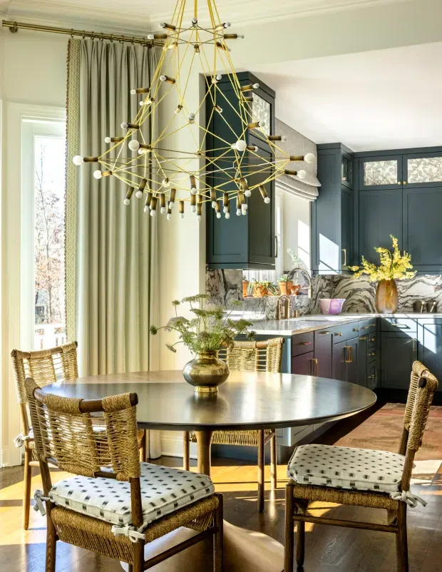 A bright, refined open-plan dining and kitchen interior. A round dark dining table with woven rattan dining chairs occupies the foreground beneath an oversized sculptural brass-and-glass chandelier. Behind it is a dark blue-grey kitchen with shaker-style cabinetry, brass hardware, a veined stone backsplash and benchtop, and a central island. Full-height curtains, classic wall mouldings, timber flooring, and abundant natural light create a sophisticated transitional setting.