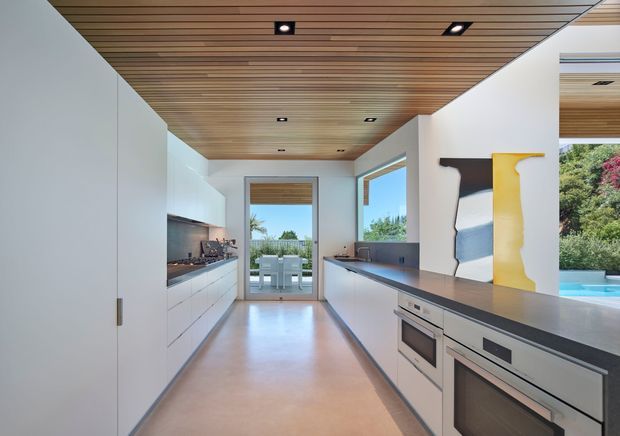 A bright, elongated contemporary kitchen with white full-height cabinetry on both sides, a dark grey stone island benchtop with integrated ovens, and a warm timber slatted ceiling with recessed lighting. Large glazed openings connect the kitchen to a dining terrace and a landscaped outdoor area with a swimming pool visible beyond. Abstract artwork adds a small accent of colour.