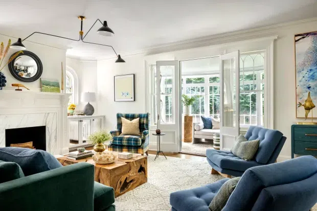 Bright, refined living room with a white traditional architectural shell, ornate fireplace mantel, detailed cornices, tall multi-pane windows and French doors opening to a conservatory or outdoor area. The room is furnished with deep blue and teal upholstered seating, a rustic live-edge timber coffee table, patterned rug, art, plants and sculptural lighting.