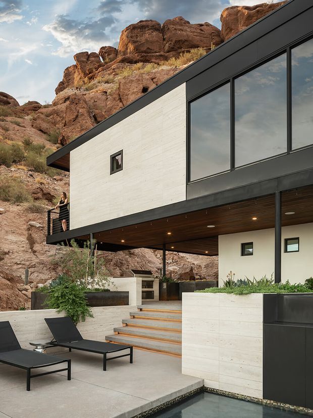 Exterior view of a modern multi-level residence built directly into a rugged red-rock hillside. The house features a sharply sloping dark roofline, large black-framed glazed openings, pale concrete or stone wall surfaces, a timber-lined soffit with recessed lighting, cantilevered upper-level massing, and a landscaped terrace with black loungers and a narrow reflecting pool. The setting is rocky and mountainous, with the architecture integrated into the steep site.