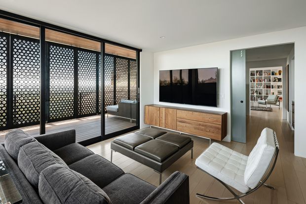 Contemporary living room with a large charcoal sectional sofa, black leather ottoman, and white lounge chair arranged around a wall-mounted television. A timber sideboard provides warm contrast against white walls, while full-height glazed doors open to a screened outdoor terrace with patterned privacy panels. A doorway leads to a bookshelf-lined adjoining space.