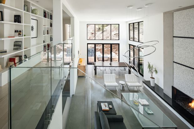 Bright, refined contemporary living room viewed from an elevated mezzanine or landing. The double-height space features extensive floor-to-ceiling glazing, a glass balustrade, white built-in shelving, a sculptural modern pendant, pale lounge furniture, dark seating, and a linear fireplace set into a textured feature wall. A lower-level living area is visible beneath the open void.