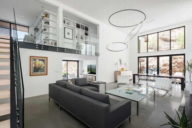 Bright open-plan living and dining area with a double-height ceiling, polished concrete flooring, large black-framed windows, and a mezzanine-level library or study overlooking the room. A dark sectional sofa faces a glass coffee table, while a glass dining table with modern chairs sits near the glazing. Sculptural circular pendant lights and white built-in shelving create a contemporary, gallery-like interior.