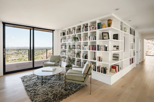 Bright contemporary living room with floor-to-ceiling black-framed glazing opening to a balcony, a panoramic city and landscape view, pale timber flooring, a large textured rug, lounge chairs and coffee table, and extensive white built-in open shelving filled with books and decorative objects. A hallway extends beyond the custom shelving.