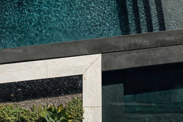 Close-up view of a contemporary pool edge and adjoining hardscape. Clear blue-green water occupies the upper left, bordered by dark charcoal coping and a pale stone-paved corner. A narrow black pebble or aggregate strip and planted greenery appear along the lower edge, with submerged pool steps visible on the right. The composition emphasizes refined pool detailing, contrasting textures, and geometric lines.