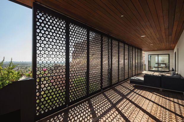 Covered outdoor terrace with a long black geometric perforated privacy screen, warm timber-lined ceiling, timber decking, and dark built-in or outdoor lounge seating. Strong patterned sunlight and shadows fall across the deck, with a broad suburban landscape visible beyond the screen.