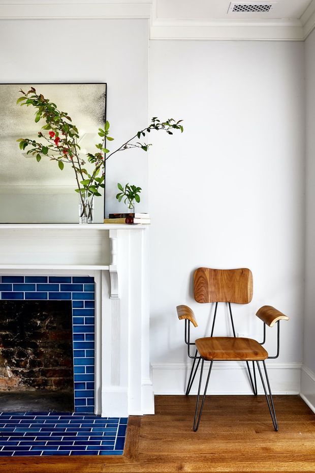 Bright, refined interior corner featuring a traditional white-painted fireplace mantel with decorative mouldings and a vivid navy-blue glazed brick-tile surround and hearth. A vintage-style wooden armchair with black metal legs sits on warm timber flooring, while a large mirror, leafy branches, and small decorative objects rest on the mantel. White walls, ornate ceiling trim, and a ceiling vent complete the room.