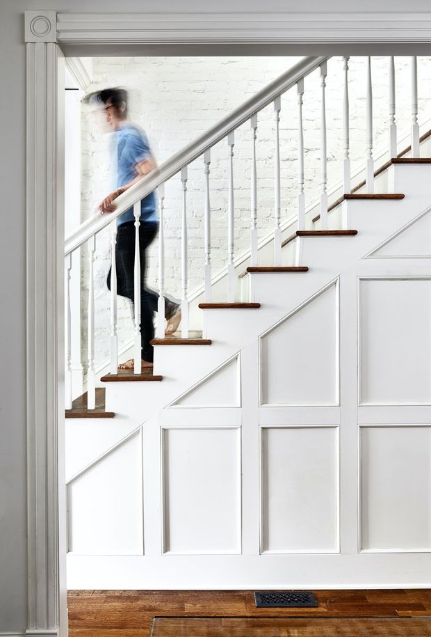 Bright interior view through a detailed white doorway or opening toward a staircase. The staircase has white painted risers and balustrade, dark timber treads, turned spindles, and decorative paneled wainscoting beneath. A motion-blurred person is ascending the stairs. Warm timber flooring and white-painted brick walls create a high-contrast traditional interior.