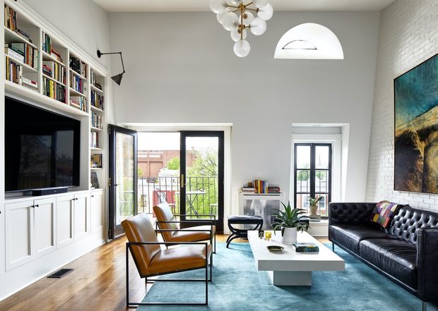 A bright, high-ceilinged living room with white built-in bookcases and TV cabinetry along one wall, large glazed doors opening to a balcony, and a tall window. The room features warm timber flooring, a large blue rug, a black tufted leather sofa, two cognac leather lounge chairs, a white rectangular coffee table, contemporary artwork, and a sculptural globe chandelier. White painted brick is visible on the right wall, with an arched clerestory window above.