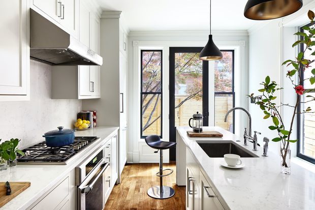 Bright, narrow galley-style kitchen with white shaker cabinetry, pale stone worktops, integrated stainless-steel appliances, a gas cooktop, and a central island with sink and raised bar seating. Tall black-framed glazed doors and windows bring in abundant daylight, while black pendant lights and natural timber flooring add contrast. The room has traditional architectural detailing paired with contemporary fixtures and restrained styling.