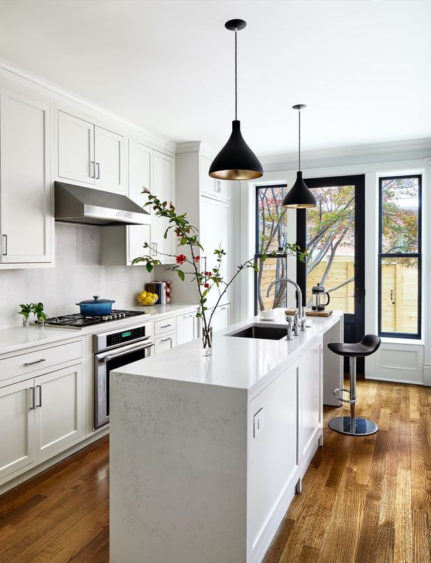 Bright, refined kitchen with white Shaker-style cabinetry, a large white stone island with integrated sink, stainless-steel appliances, and warm timber flooring. Two black pendant lights hang above the island, while black-framed glazed doors and windows open to a fenced outdoor area. The room combines classic detailing with contemporary fixtures and a high-contrast black-and-white palette.