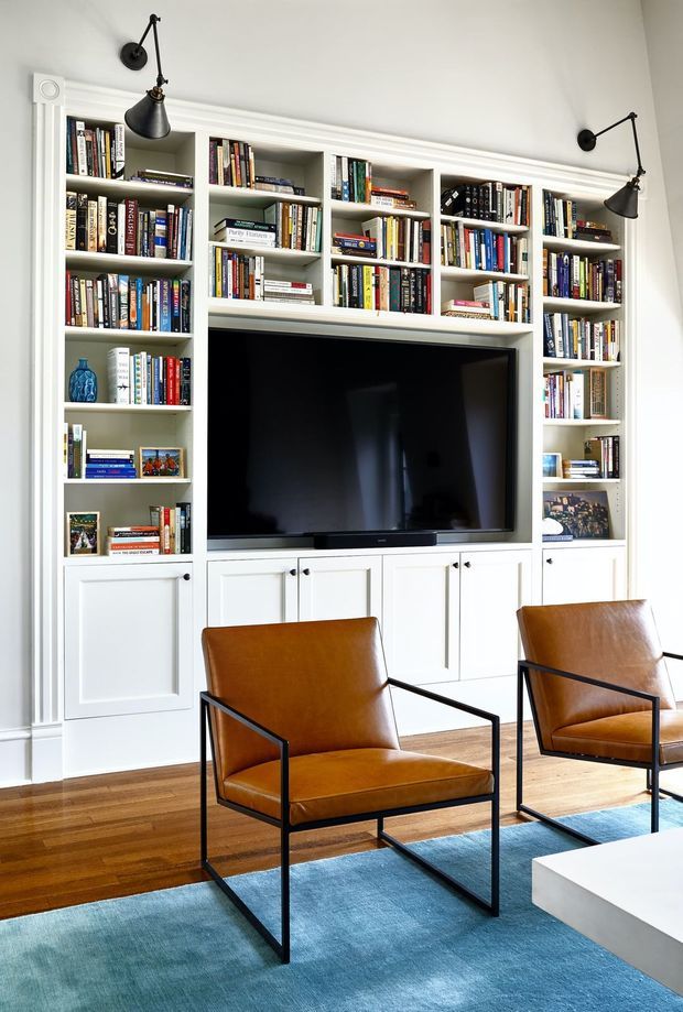 Bright, refined living room featuring a full wall of custom white built-in bookcases with integrated lower cabinets and a large flat-screen television. The shelving is filled with books and decor, and two cognac leather armchairs with slim black metal frames sit on a blue area rug over warm timber flooring. Black articulated wall lights flank the shelving, while classic white moulded trim adds architectural detail.