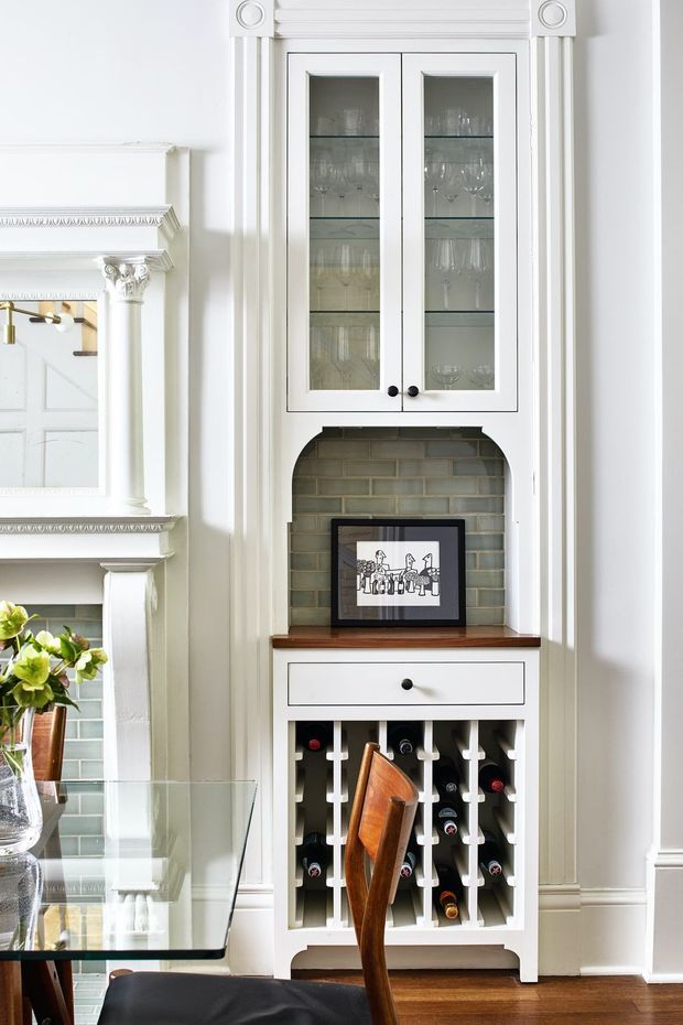 A bright, traditionally detailed dining area featuring a custom white built-in wine cabinet and display hutch. The upper glazed doors display glassware, while the lower open section stores wine bottles beneath a white drawer and dark timber benchtop. The cabinet is set within ornate white wall panelling beside a classic fireplace surround, with a glass dining table and dark timber flooring visible in the foreground.