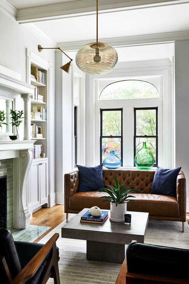 Bright, refined sitting room with a traditional fireplace and extensive white built-in bookshelves, a tufted brown leather sofa, dark blue cushions, and a substantial concrete coffee table. White decorative wall panelling and trim frame black-painted windows with an arched transom, while a sculptural glass pendant and brass articulated wall lamp provide warm accents. Timber parquet flooring and a large neutral rug complete the layered interior.