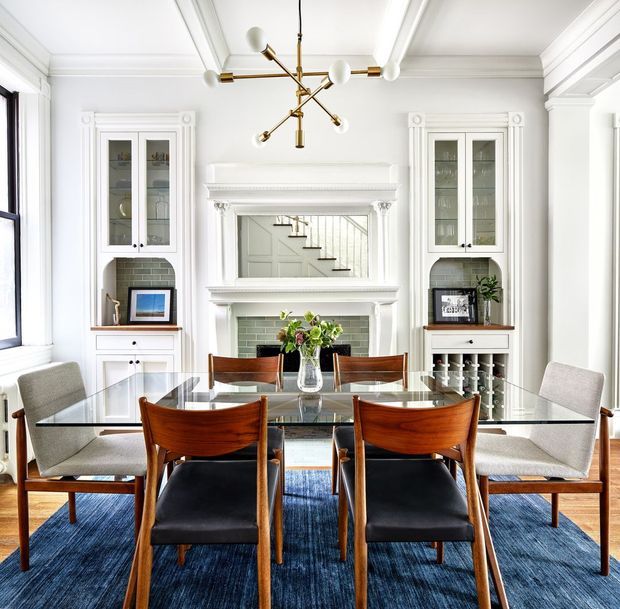 Bright, refined dining room with a glass-top dining table surrounded by mid-century-style wooden and upholstered chairs. The room features a decorative white fireplace with green tiled surrounds, symmetrical built-in display cabinetry, detailed wall and ceiling mouldings, a large mirror, and a sculptural brass-and-white pendant light. Blue area rug and dark upholstered seating add contrast to the predominantly white interior.