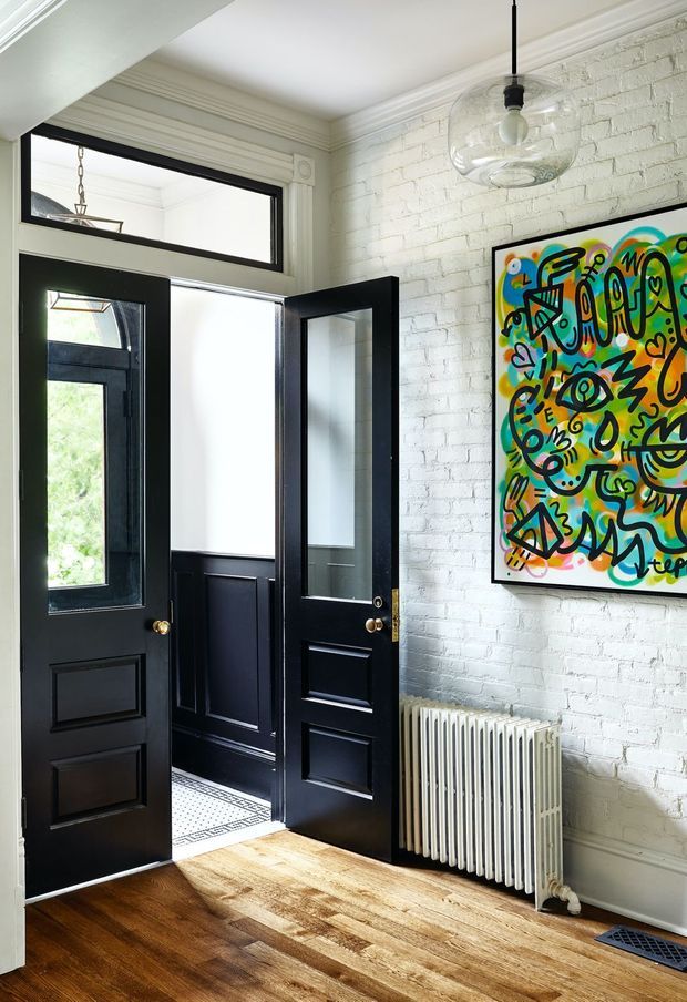 Bright traditional entryway featuring polished dark timber flooring, black paneled double doors with glazed upper sections, white-painted exposed brick walls, ornate ceiling cornices, and a high transom window. A vintage-style radiator and colourful framed artwork add character, while a pendant light hangs from the ceiling. The open doors reveal a small adjoining hall with dark wainscoting and patterned tile flooring.