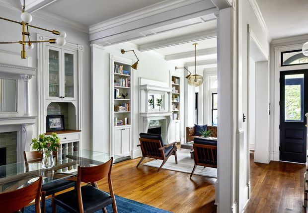 Bright, refined interior view of a traditional home showing a dining area in the foreground opening into a living room. The rooms feature polished timber flooring, white painted wall panelling and detailed mouldings, built-in shelving, a fireplace surround, black-framed glazing, and sculptural contemporary lighting. Rich wood furniture and a blue rug add contrast to the predominantly white interior.