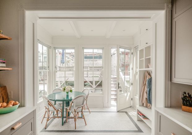 Bright, narrow enclosed dining sunroom opening to an exterior deck or porch. The room features white-painted architectural trim and built-in cabinetry, a glazed bay of windows and a glazed door, a small round glass dining table with wicker chairs, and a patterned light floor bordered in grey. A coat storage nook and shelving are visible on the right, with kitchen-style joinery framing the space.