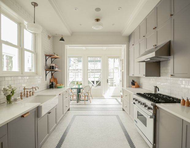 Bright, spacious galley-style kitchen with pale grey shaker cabinetry, white subway-tile splashbacks, white stone-look benchtops, and a large freestanding stainless-steel range cooker. A patterned light floor runner leads toward a sunlit dining nook with French doors and windows. Traditional trim, ceiling mouldings, pendant lighting, and brass hardware create a refined classic interior.