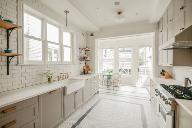 Bright, elongated kitchen with white subway-tile walls, pale grey shaker cabinetry, white countertops, a farmhouse-style sink, and a large stainless-steel range. Tall windows and glazed double doors at the far end flood the space with natural light and open toward a small outdoor area. Open timber shelving, brass-toned hardware, and a patterned mosaic floor add detail.