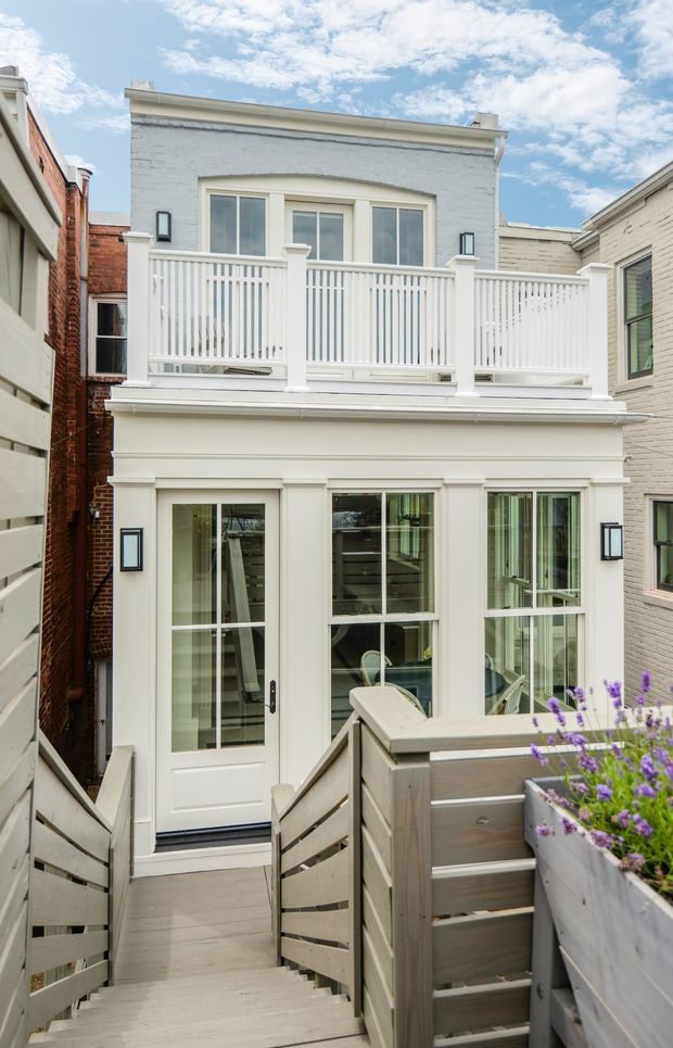 A narrow urban residential exterior viewed from a raised timber deck and stairway. The compact two-storey facade is painted white with textured masonry walls, classical trim around large glazed doors and windows, and a white-railed upper balcony. Timber privacy screens, planter boxes with purple flowers, and neighbouring attached buildings frame the courtyard-like setting.