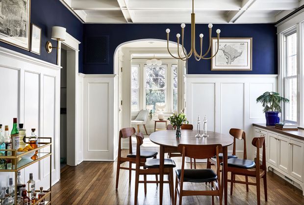 A refined dining room with a round wooden dining table and mid-century-style wooden chairs, set against deep navy upper walls and white decorative wainscoting. The room features a white coffered ceiling, brass chandelier and wall sconces, dark timber flooring, built-in cabinetry, framed artwork, and an arched opening leading to a bright adjoining sitting area. A bar cart and indoor plant add functional and decorative details.