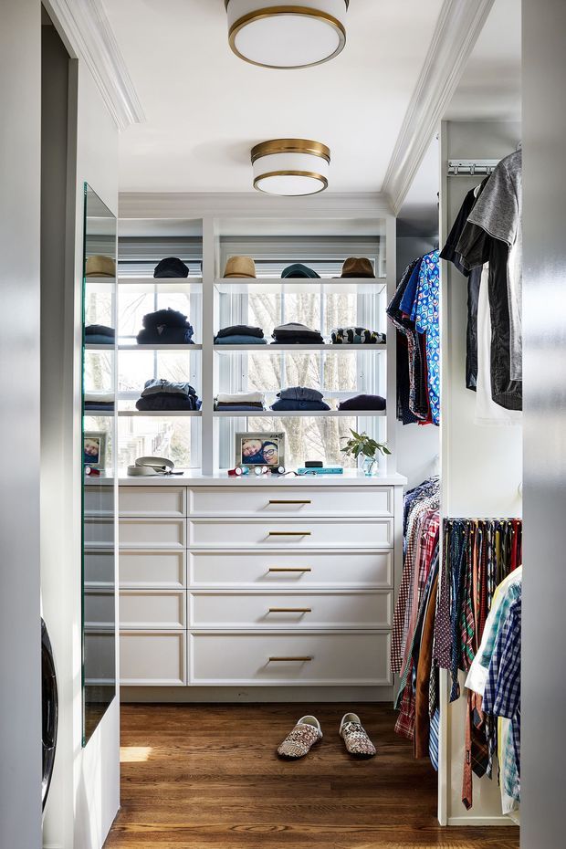 A bright, narrow walk-in wardrobe with extensive custom white built-in cabinetry, open shelving, drawers, hanging rails and mirrored side panels. Clothing, hats and accessories are neatly displayed, with warm timber flooring, white architectural trim, and brass-and-white flush-mount ceiling lights. Large windows at the rear provide abundant natural light.