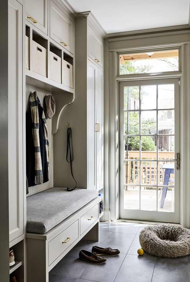 A bright, built-in mudroom entryway with full-height painted cabinetry, open cubbies containing storage baskets, a coat area, upholstered bench seat, and drawers with brass pulls. A glazed exterior door with transom window provides daylight and opens to a small deck. Large grey floor tiles, detailed trim, and a pet bed reinforce the practical, refined character of the space.