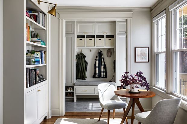 A bright, refined entryway or mudroom nook with built-in white cabinetry, open shelving filled with books and objects, overhead storage bins, coat hooks, and a built-in bench with drawers. A small round timber table, upholstered chairs, brass wall light, framed artwork, and large windows complete the warm traditional interior.