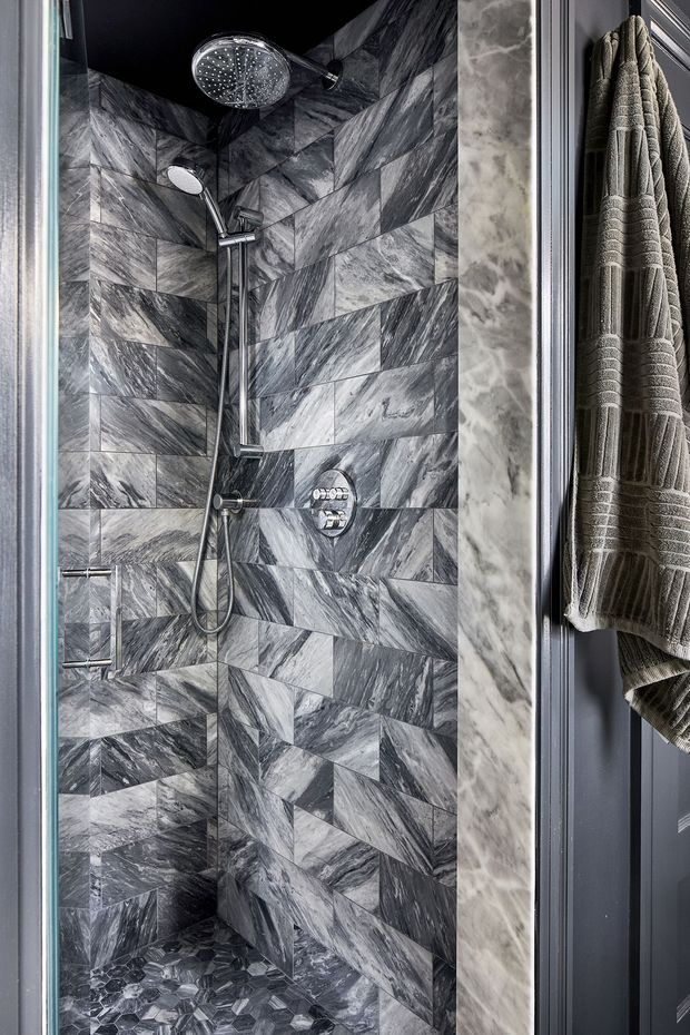 A compact, luxurious walk-in shower enclosed within a bathroom. The walls and floor are clad in dramatic veined grey marble-look tiles, with a contrasting dark ceiling, chrome shower controls, a large rain showerhead, handheld shower, and built-in niche seating. A textured grey towel hangs on a dark paneled door or wall beside the shower.