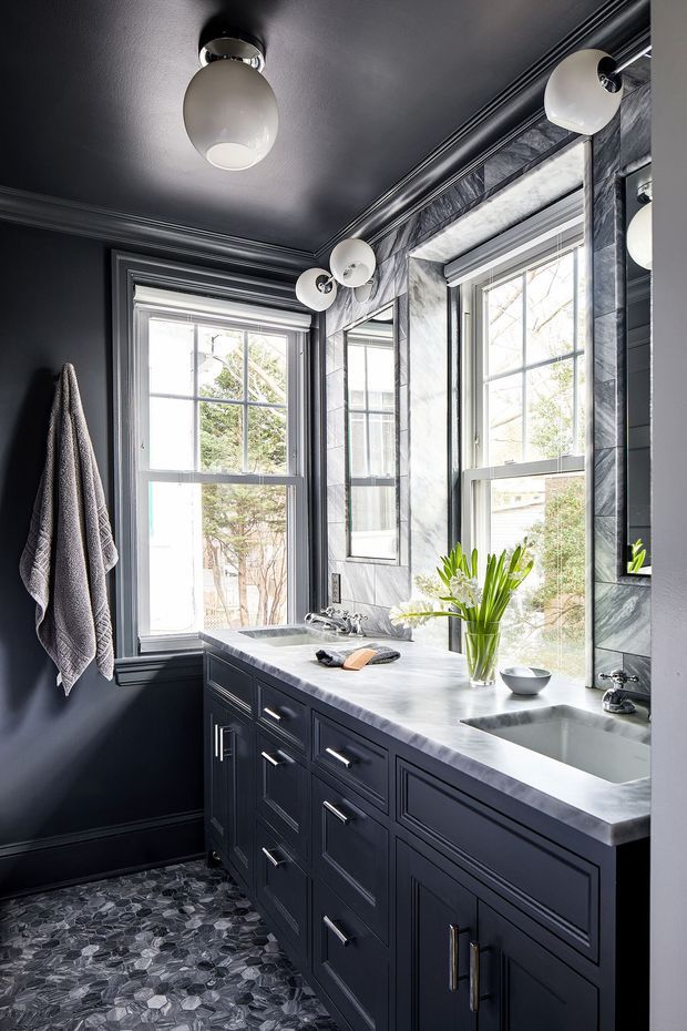 A compact, elegant bathroom with a dark charcoal palette, extensive framed windows, a long dark built-in vanity, pale stone countertop, integrated basin, and large mirror. The walls and ceiling feature traditional decorative trim, while globe wall and ceiling lights provide a refined statement. The floor is finished with a patterned dark stone tile, and a towel hangs beside the window.