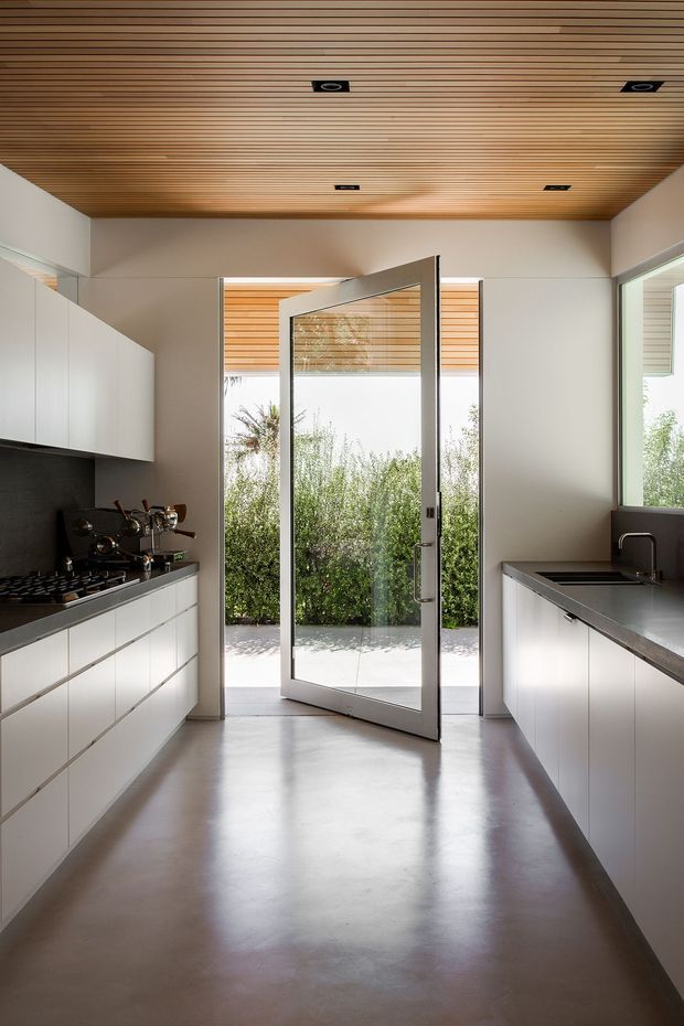 A clean, contemporary galley kitchen viewed toward an open glazed exterior door. The room features white flat-panel cabinetry on both sides, dark charcoal benchtops and splashback, integrated cooking and sink areas, a smooth polished grey floor, and a warm slatted timber ceiling with recessed downlights. Bright greenery is visible outside through the doorway and side glazing.