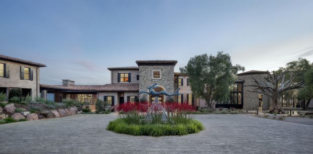 A broad exterior view of a low-rise, multi-building residential or hospitality-style courtyard arranged around a large paved forecourt. The architecture features stucco walls, dark timber or metal window frames, tiled pitched roofs, stone accents, and a central tower-like volume with a decorative sculptural fountain and planting. Mature trees, landscaped rock beds, and ornamental grasses soften the expansive grey paving.
