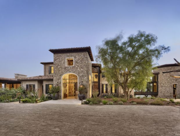 Exterior view of a sprawling low-rise residence with a central two-storey stone entry tower, arched wrought-metal gate, warm timber-framed windows, and broad tiled rooflines. The building is arranged around landscaped planting and a large paved forecourt, with a warm Mediterranean or rustic villa character.