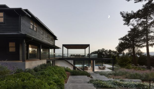 Exterior view of a contemporary multi-level residence set within landscaped grounds. The dark horizontal-clad house has broad glazing, projecting roof planes, a glass-balustraded terrace, and a lower-level outdoor living area with a framed pavilion and stairs. Mature trees and distant terrain surround the property in soft evening light.