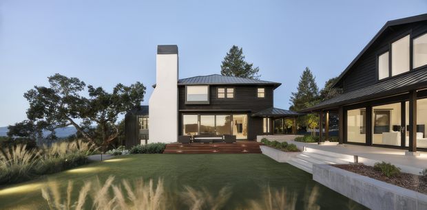 A contemporary multi-wing residence viewed from a landscaped courtyard. The home features dark metal-clad walls and pitched roofs, expansive floor-to-ceiling glazing, a tall pale chimney, timber decking and steps, and a broad lawn with ornamental grasses. A second connected or adjacent wing frames the right side of the composition, while mature trees and a distant blue horizon form the background.
