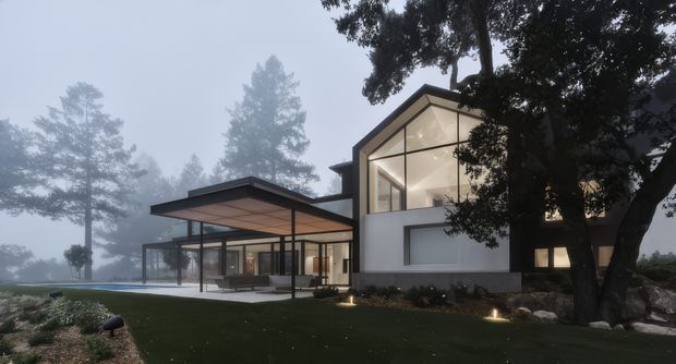 Exterior view of a contemporary multi-level residence in a misty, wooded setting. The house features a strong geometric composition, flat and low-pitched roof planes, extensive floor-to-ceiling glazing, a projecting covered outdoor terrace, white rendered walls, and dark structural framing. Landscape lighting and mature trees frame the facade.