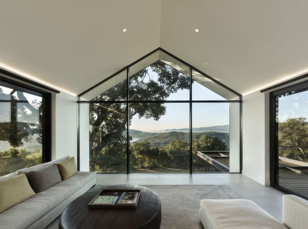 A bright contemporary living room with expansive black-framed floor-to-ceiling glazing forming a peaked central window wall, offering views of a wooded sloping landscape and distant hills. The room has white walls and ceiling with recessed spotlights and concealed perimeter lighting, pale tiled flooring, a large light-grey sectional sofa, a rounded dark coffee table, and a pale upholstered ottoman. The composition emphasizes the strong indoor-outdoor connection and panoramic outlook.