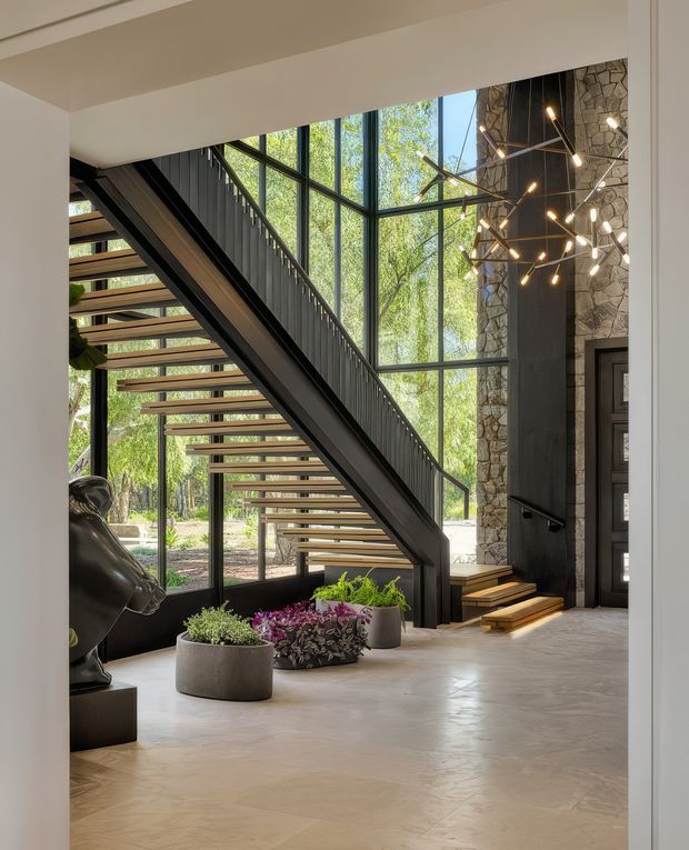 A bright, sophisticated residential entry hall featuring a dramatic open-riser staircase with dark metal stringers and balustrade, timber treads, expansive black-framed glazing, and a tall natural stone feature wall. A sculptural pendant chandelier hangs beside the staircase, while planted pots and a sculptural figure add visual interest to the polished pale floor.