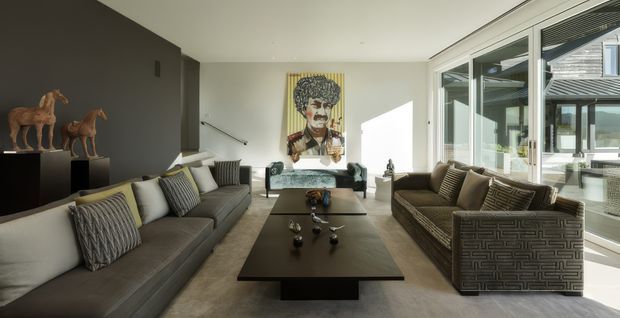 A bright, spacious contemporary living room with two large sofas arranged around a long dark coffee table. The room features a dark charcoal feature wall with sculptural horse objects, neutral upholstered seating with patterned cushions, a pale textured floor, and expansive glazed corner windows opening visually toward an outdoor courtyard or terrace. A colourful artwork provides a focal point, while strong daylight creates a refined, gallery-like atmosphere.