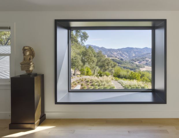 Interior view centered on a large deep-set black-framed picture window, presenting a panoramic mountainous landscape. The room has smooth light walls, pale timber flooring, and a dark sculptural display pedestal with artwork at the left. The composition is minimal and gallery-like, with strong natural light and a clear connection to the surrounding landscape.