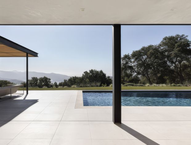 A minimalist covered outdoor terrace overlooking a rectangular swimming pool and open landscaped grounds. Large white tiled paving, slim dark structural posts, and a flat roof create a clean contemporary setting, with distant hills and mature trees visible beyond the pool.